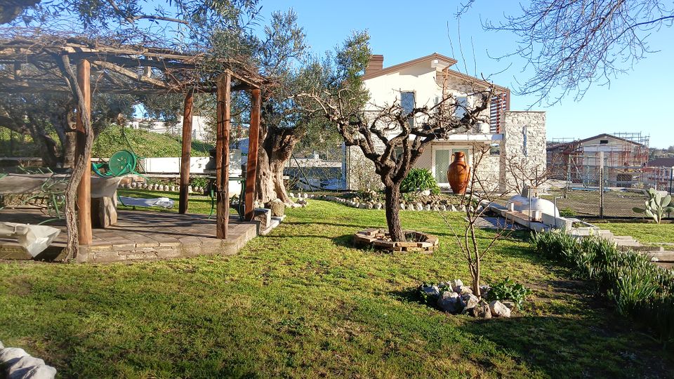 Cortile di una casa di campagna con prato e alberi, con pergolato in legno e arredi da giardino sulla sinistra. Sullo sfondo si vede un’abitazione a due piani con facciata in pietra e balcone, oltre a piccoli muretti in pietra e aiuole.
