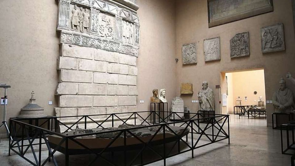 Sala museale con pareti color sabbia che espone un grande rilievo lapideo monumentale con fregi scolpiti, affiancato da statue e pannelli in pietra. Al centro c’è una struttura rettangolare recintata in metallo e, sul fondo, un passaggio aperto conduce a un’altra galleria.