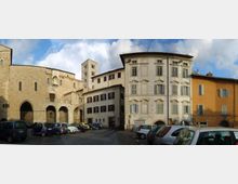 Piazza urbana con parcheggio di auto, circondata da edifici storici: una chiesa in pietra con archi e portali a sinistra e palazzi residenziali a più piani con facciate intonacate e finestre regolari a destra. Sullo sfondo si vede un campanile in pietra sotto un cielo parzialmente nuvoloso.