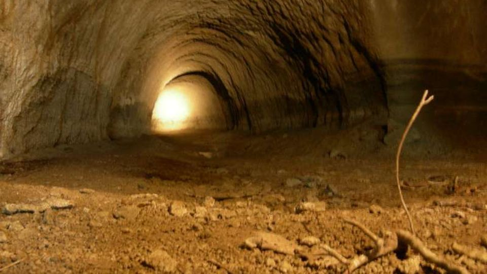Interno di un tunnel sotterraneo con pareti e soffitto in terra compatta, illuminato da una luce intensa visibile in fondo. Il pavimento è polveroso e irregolare, con piccoli sassi e rami secchi in primo piano.