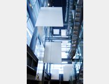 Atrio interno di un edificio moderno con pareti vetrate e struttura in acciaio, illuminato da luce naturale dall’alto. Grandi pannelli bianchi sospesi al soffitto scendono verticalmente nello spazio centrale sopra passerelle e scale in vetro.