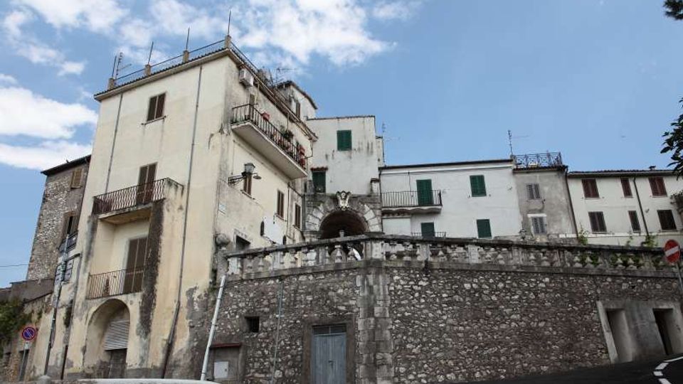 Edifici residenziali a più piani su un pendio, con facciate intonacate chiare, persiane verdi e balconi in ferro. In primo piano un alto muro di contenimento in pietra con parapetto e una strada asfaltata curva sotto un cielo parzialmente nuvoloso.