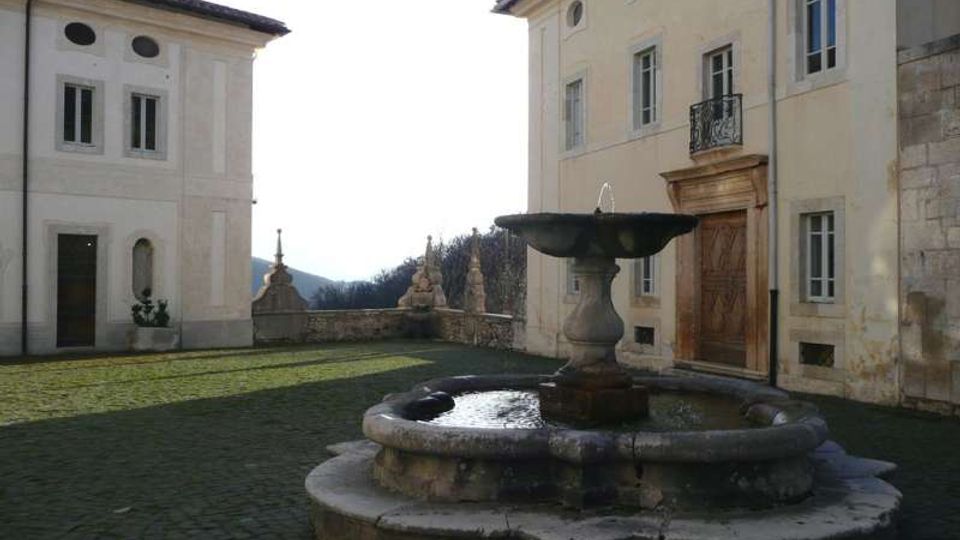 Cortile lastricato con una fontana in pietra al centro, circondato da edifici storici intonacati con finestre alte, porta monumentale e piccolo balcone in ferro battuto. Sullo sfondo si vede un muretto con elementi decorativi e un paesaggio collinare.