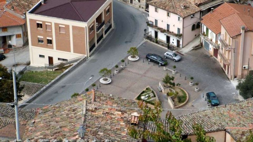 Veduta dall’alto di una piccola piazza di paese con parcheggio, aiuole e panchine, circondata da edifici a più piani con facciate chiare e tetti in tegole. Una strada curva costeggia la piazza e collega le case del centro storico.