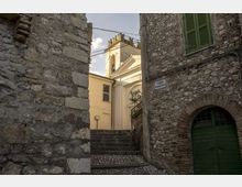 Vicolo in pietra con scalinata e pavimentazione in ciottoli tra edifici in muratura, con arco e portone verde e finestra con persiane. In fondo si intravede la facciata chiara di una chiesa con campanile e cavi sospesi sopra la strada.