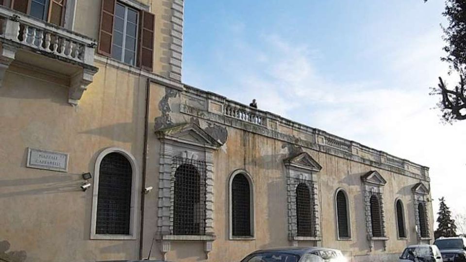 Facciata di un edificio storico in intonaco giallo con finestre ad arco e grandi grate in ferro, cornici in pietra e piccoli frontoni sopra alcune aperture. Davanti si vede una strada con auto e motocicli parcheggiati, sotto un cielo sereno e con alberi sul lato destro.