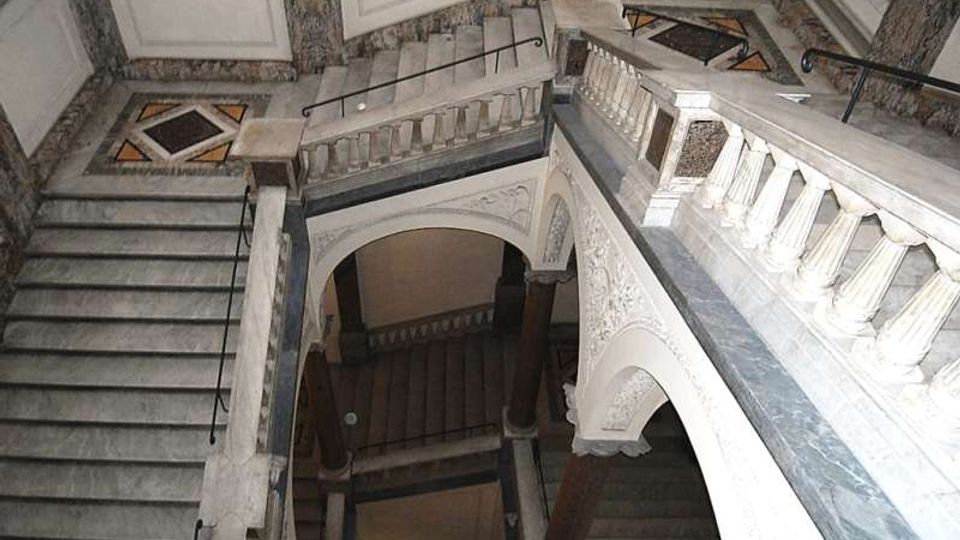 Interno di un vano scala monumentale in marmo con rampe sovrapposte, balaustre scolpite e ampi archi decorati. Pareti rivestite in pietra e pavimenti a motivi geometrici con corrimani in metallo lungo i gradini.