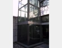 Angolo di un edificio moderno con struttura in metallo scuro e grandi pannelli di vetro, simile a un vano ascensore o ingresso vetrato. Accanto si vede un muro rivestito in pietre a mosaico e una pavimentazione esterna in pietra, con ombre di alberi riflesse sulle superfici.