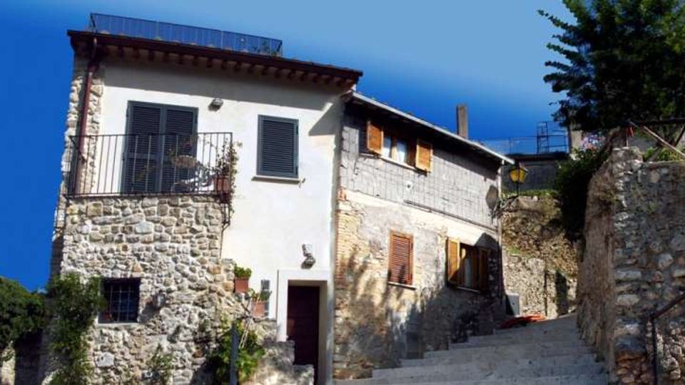 Edifici in pietra e intonaco bianco su una strada in salita, con persiane scure, balcone con ringhiera in ferro e finestre con scuri in legno. Il contesto è un borgo collinare con muri in pietra e vegetazione ai lati sotto un cielo limpido.