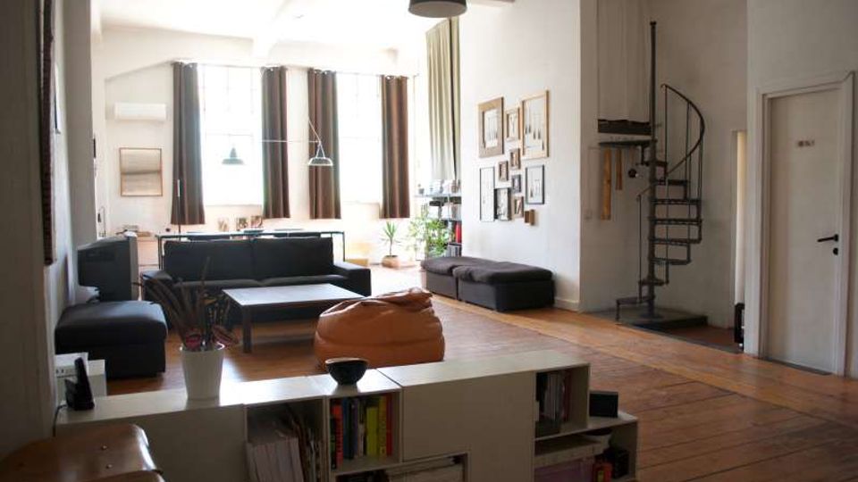 Soggiorno open space in stile loft con pavimento in legno, grandi finestre con tende scure e arredi moderni (divano, tavolino e pouf). Sulla destra una scala a chiocciola in metallo conduce a un soppalco, con pareti bianche decorate da una composizione di quadri.