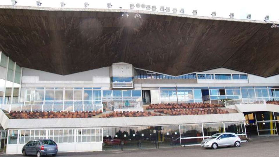 Facciata di una tribuna di impianto sportivo con grande tetto sporgente in legno, ampie vetrate su più livelli e file di sedili visibili. Davanti si trova un piazzale asfaltato con alcune auto parcheggiate.