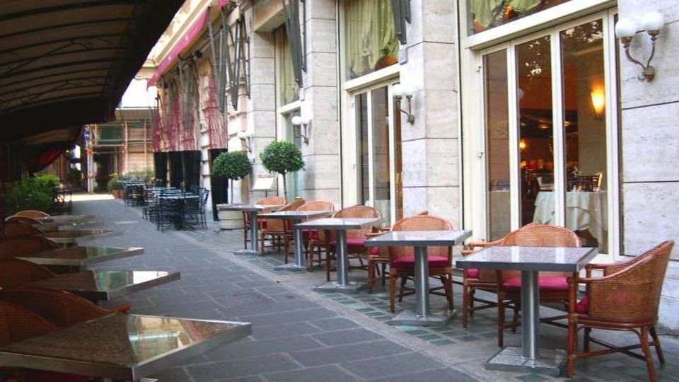 Dehors di un ristorante lungo un portico coperto, con file di tavoli e sedie in rattan su pavimentazione in pietra. Sul lato destro si vede la facciata in pietra con grandi vetrate e tende, illuminate da applique a parete.