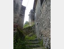 Vicolo stretto in un borgo antico con scalini in pietra invasi da erba e vegetazione, affiancato da alte pareti di case in pietra grezza e intonaco. Sullo sfondo si vedono muri a secco e un edificio con tetto sporgente e tubazioni esterne.