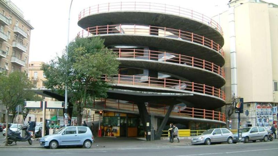 Edificio urbano a pianta circolare con più livelli sovrapposti e rampe elicoidali esterne, evidenziate da ringhiere rosse, affacciato su una strada trafficata. Alla base si vedono pilastri di sostegno, un’area d’ingresso e alberi e auto parcheggiate lungo il marciapiede.