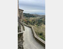 Veduta da un borgo collinare con una strada pedonale in pietra in discesa, affiancata da muri e ringhiere, che costeggia edifici in muratura. Sullo sfondo si apre una valle boscosa con campi e colline lontane sotto un cielo nuvoloso.