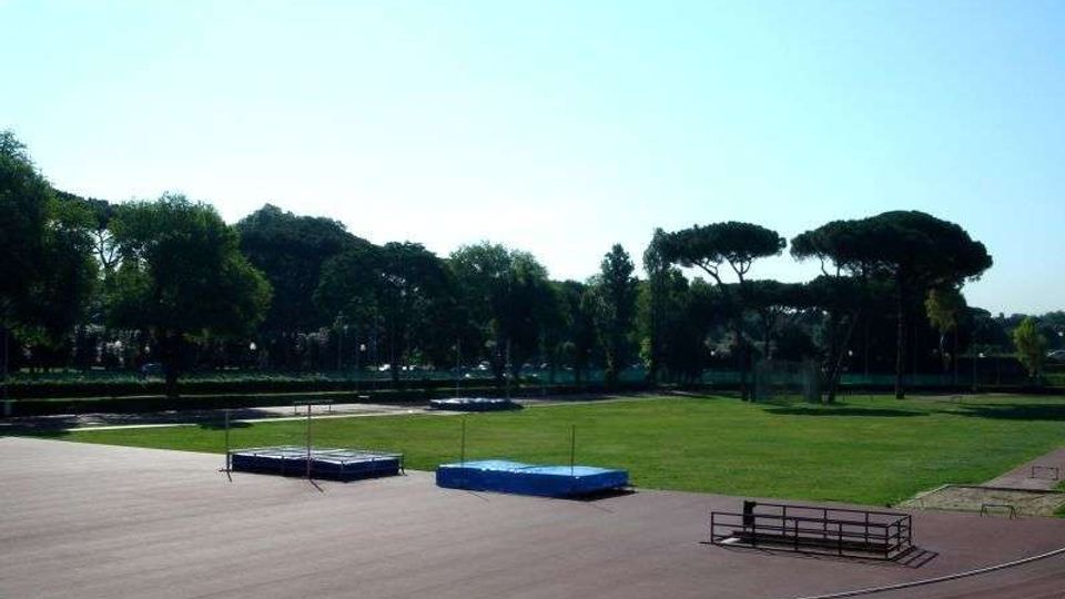 Pista di atletica all’aperto con ampio prato centrale e pedane per salto in alto con materassi blu. Sullo sfondo si vedono alberi alti e una linea di verde che delimita l’area sportiva.