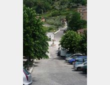 Piazza di un piccolo paese con pavimentazione in pietra, parcheggi laterali e una fontana al centro vicino a un edificio bianco. Sullo sfondo una strada sale verso case sparse su una collina verde con alberi e uliveti.