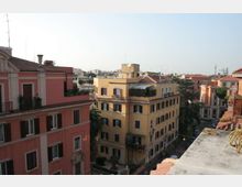 Veduta urbana dall’alto con palazzi residenziali in stile tradizionale dai toni rosa e giallo, con tetti in tegole, balconi e finestre con persiane. Sullo sfondo si vede un quartiere fitto di edifici e una strada alberata tra i palazzi.