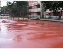 Campo sportivo all’aperto con pavimentazione rossa bagnata dalla pioggia e recinzione metallica alta lungo il perimetro. Sullo sfondo si vedono alberi e un edificio residenziale con balconi e finestre.