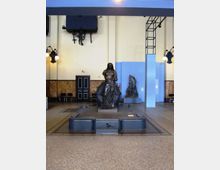 Sala interna in stile industriale con pavimento a mosaico e pareti chiare con pannelli blu; al centro una grande scultura in bronzo su basamento, circondata da una struttura metallica incassata nel pavimento. Sullo sfondo si vedono una porta, sedute laterali, lampade a parete e un secondo rilievo esposto su piedistallo.
