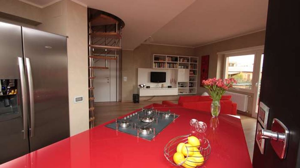 Interno di un appartamento open space con cucina moderna: grande isola rossa lucida con piano cottura integrato e frigorifero in acciaio. Sullo sfondo si vede il soggiorno con divano rosso, parete attrezzata con librerie e TV, scala a chiocciola e una finestra che illumina l’ambiente.