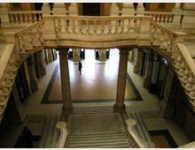 Atrio interno di un edificio storico con ampie scale in pietra e balaustre scolpite che si affacciano su un pianerottolo inferiore con colonne e pavimento a motivi geometrici. Ai lati si vedono corridoi con porte e, sul fondo, un’ampia apertura verso l’esterno.