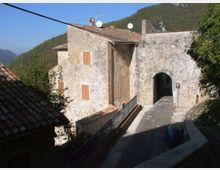 Edificio in pietra e intonaco con tetto in coppi e finestre con persiane, affiancato a un grande arco che forma un passaggio coperto su una strada in salita. Sullo sfondo si vedono colline boscose e cielo sereno.