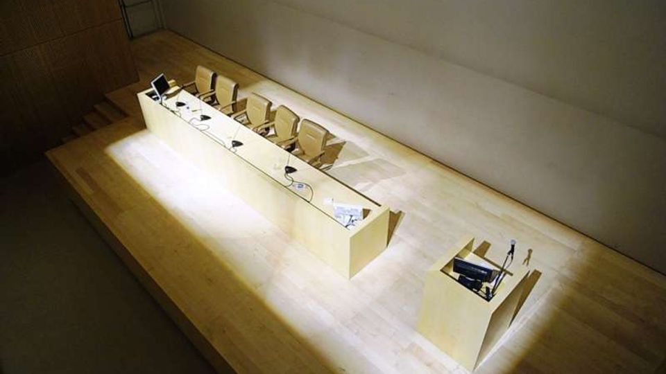 Sala conferenze/auditorium con palco in legno chiaro, lungo tavolo per relatori con sedie, microfoni e monitor, illuminato da un fascio di luce. A lato del palco c’è un piccolo podio con microfono, con pareti spoglie e un accesso a gradini.