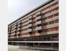 Edificio residenziale moderno a più piani, lungo e lineare, con file di balconi e pannelli/fasce rosse sulle facciate. Al livello strada si vede un porticato continuo e una recinzione metallica lungo il marciapiede.