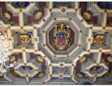 Soffitto a cassettoni riccamente decorato con riquadri geometrici, inserti dorati e pannelli blu e bordeaux. Al centro spicca uno stemma araldico entro una cornice ottagonale, con un lampadario visibile sul bordo sinistro.