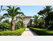 Viale di ghiaia in un giardino formale, fiancheggiato da siepi potate e alte palme, che conduce a una villa storica con facciata in pietra, finestre regolari e portale ad arco. Sullo sfondo si vede il cielo sereno e una vegetazione mediterranea densa ai lati del percorso.