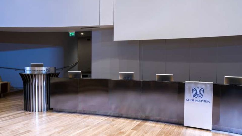Sala conferenze moderna con ampio palco in legno e lungo banco relatori in metallo, con postazioni dotate di schermi. Ai lati sono presenti due podi, uno con il logo “Confindustria”, e una parete di fondo grigia sotto un soffitto alto.