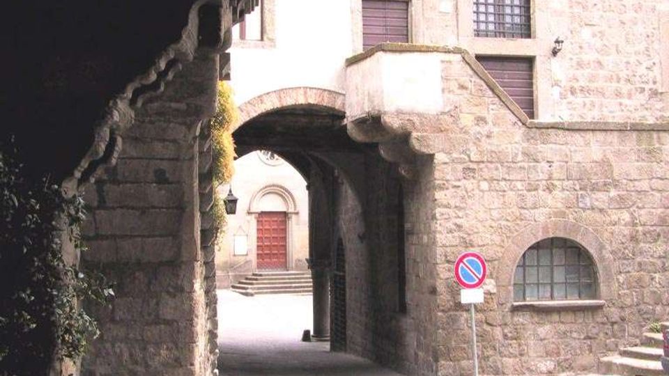Passaggio ad arco in pietra con scalinate laterali e facciate in muratura con finestre; sotto l’arco si intravede un portone rosso in fondo a una piccola piazza lastricata. Ai lati sono presenti un lampione e un segnale stradale di divieto di sosta.