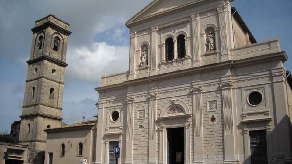 Facciata di una chiesa in stile neoclassico con frontone triangolare, colonne e tre archi superiori con statue in nicchie; a sinistra si vede un campanile in pietra a torre. Davanti all’edificio c’è una piazza con auto parcheggiate sotto un cielo parzialmente nuvoloso.