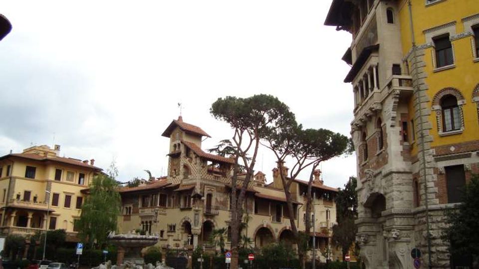 Piazza urbana con rotonda e fontana scultorea al centro, circondata da edifici storici in stile liberty con torrette e logge. Sullo sfondo si vedono alti pini marittimi e facciate in pietra e intonaco, con un palazzo giallo a destra.