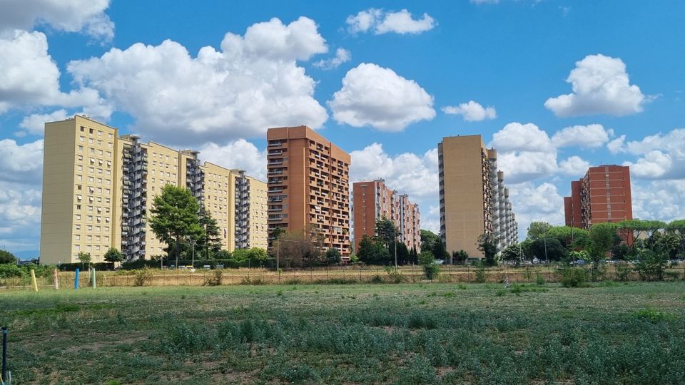 Vista di un’area aperta con vegetazione bassa in primo piano e, sullo sfondo, una fila di alti condomini residenziali in cemento e mattoni con balconi. Il cielo è azzurro con grandi nuvole e alberi lungo il margine del quartiere.