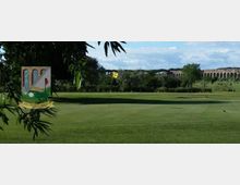 Campo da golf con green ben curato e bandierina gialla, circondato da prati e alberi. Sullo sfondo si vede un lungo acquedotto in pietra che attraversa il paesaggio sotto un cielo parzialmente nuvoloso.