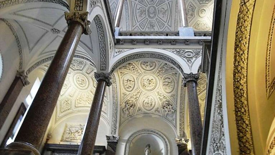 Interno di un edificio storico con grande scalone e loggiati, sorretto da alte colonne in pietra. Le volte e i soffitti sono decorati con stucchi geometrici e rosoni, con una statua in nicchia al centro del livello superiore.