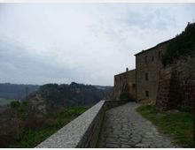Sentiero lastricato in pietra con muretto di parapetto che costeggia un edificio in muratura su un crinale. Sullo sfondo si vedono colline e una valle boscosa sotto un cielo nuvoloso.