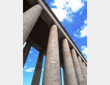 Ripresa dal basso di un viadotto in cemento armato con alte colonne cilindriche che sostengono l’impalcato superiore. Sullo sfondo si vede un cielo azzurro con nuvole sparse.