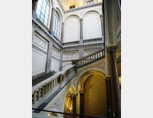 Interno di un edificio storico con ampio vano scala monumentale, pareti decorate e colonne in marmo. Grandi finestre ad arco illuminano una balconata superiore e una serie di archi e volte sottostanti.