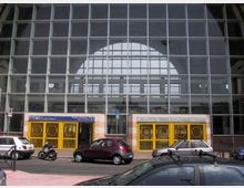 Facciata di una grande stazione ferroviaria con ampia vetrata a griglia e arco superiore, che lascia intravedere la struttura interna. Alla base ci sono ingressi con cancellate gialle e insegne, affacciati su una strada con auto parcheggiate.