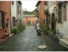 Vicolo acciottolato in un borgo storico, fiancheggiato da case intonacate con persiane in legno, lampioni a parete e numerosi vasi di piante lungo i muri. In primo piano si vedono una moto e una bicicletta parcheggiate, con una piccola piazzetta e un edificio sullo sfondo.