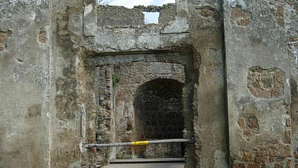 Rovine di un edificio in pietra con facciata a timpano e grande arco centrale, con muratura a vista e intonaco deteriorato. Un passaggio ad arco conduce all’interno, accessibile tramite gradini in pietra, con prato e cielo sullo sfondo.