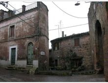Piccola piazza di borgo con edifici in muratura intonacata e facciate consumate, porte e persiane in legno e scalini in pietra. In primo piano si vedono una ringhiera bassa, cavi aerei e un grande arco in pietra sul lato destro.