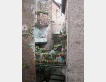 Cortile interno tra edifici in pietra e intonaco con balconi e finestre, visto attraverso un passaggio stretto. In primo piano una ringhiera con numerosi vasi di piante e, sotto, una breve scalinata che scende verso un’area inferiore.