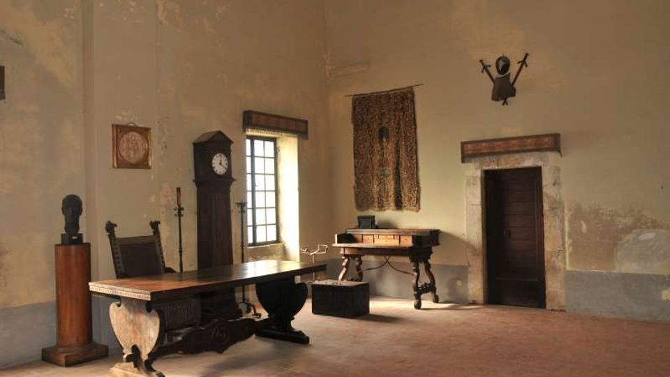 Interno di una grande stanza storica con soffitto alto e pareti intonacate, arredata con un lungo tavolo e sedie in legno scuro. Sono presenti una finestra con inferriate, un grande orologio a pendolo, un arazzo appeso, un mobile scrittoio e una porta incorniciata in pietra.