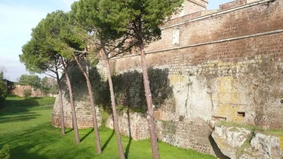 Mura di una fortezza in mattoni e pietra viste dall’esterno, con un ampio fossato erboso ai piedi. Alcuni alti pini costeggiano il muro, che presenta superfici consumate e una struttura massiccia sullo sfondo.