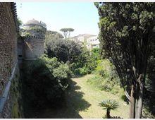 Veduta di un fossato verde con prato e vegetazione fitta ai piedi di alte mura in mattoni, con una torre cilindrica merlata sulla sinistra. Sullo sfondo si vedono alberi alti e alcuni edifici oltre il perimetro murario.