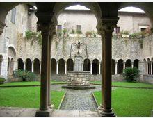 Chiostro in pietra con porticato ad archi e colonne su tre lati, affacciato su un prato centrale attraversato da un sentiero in ciottoli. Al centro si vede un pozzo in muratura con struttura in ferro, con muri in pietra e vasi di piante sul livello superiore.